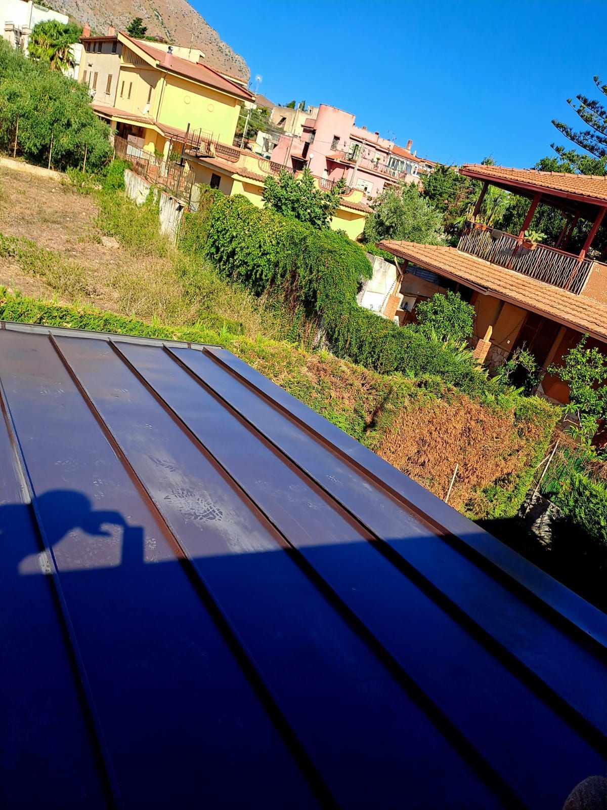 In primo piano, un tetto metallico blu si affaccia sulle case sulla collina sotto un cielo azzurro brillante.