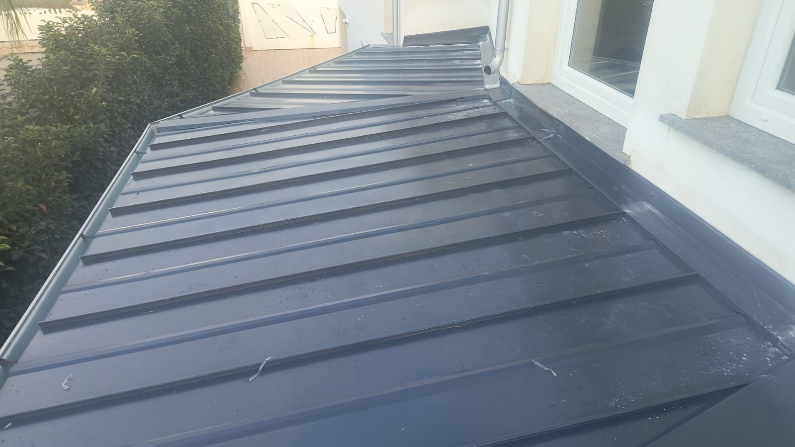 Tenda da sole in metallo grigio scuro sopra un vialetto laterale accanto a una casa bianca e cespugli verdi.