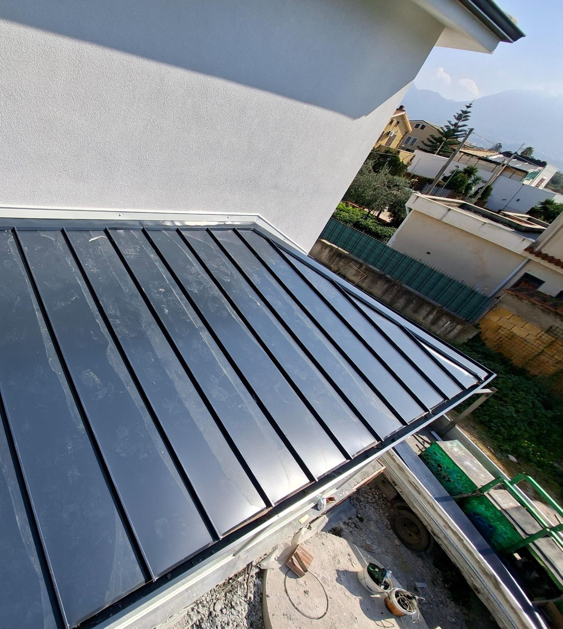 Pannelli metallici per il tetto installati su una casa accanto a un muro bianco, con un quartiere residenziale sullo sfondo.