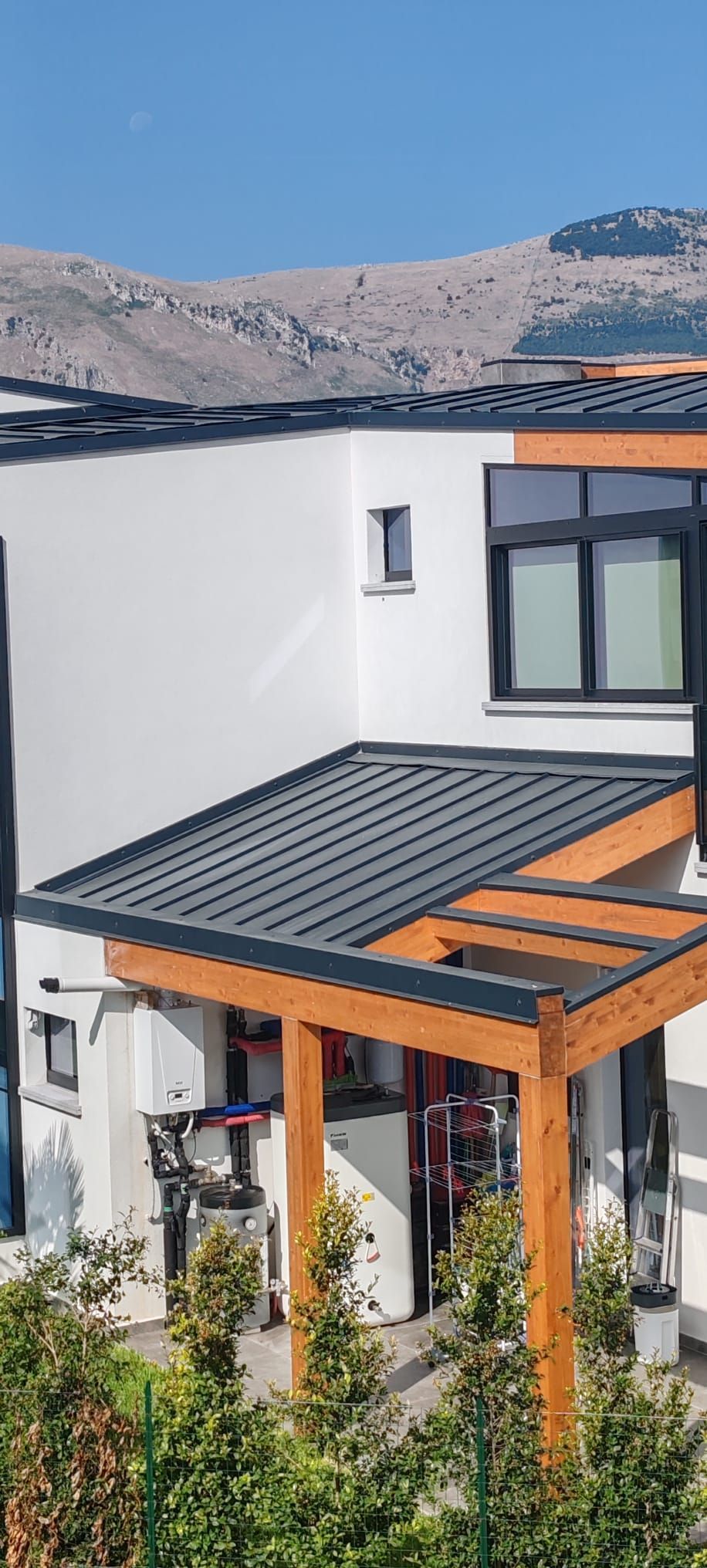 Casa moderna bianca con veranda in legno e tenda da sole nera, sullo sfondo di colline e cielo azzurro.