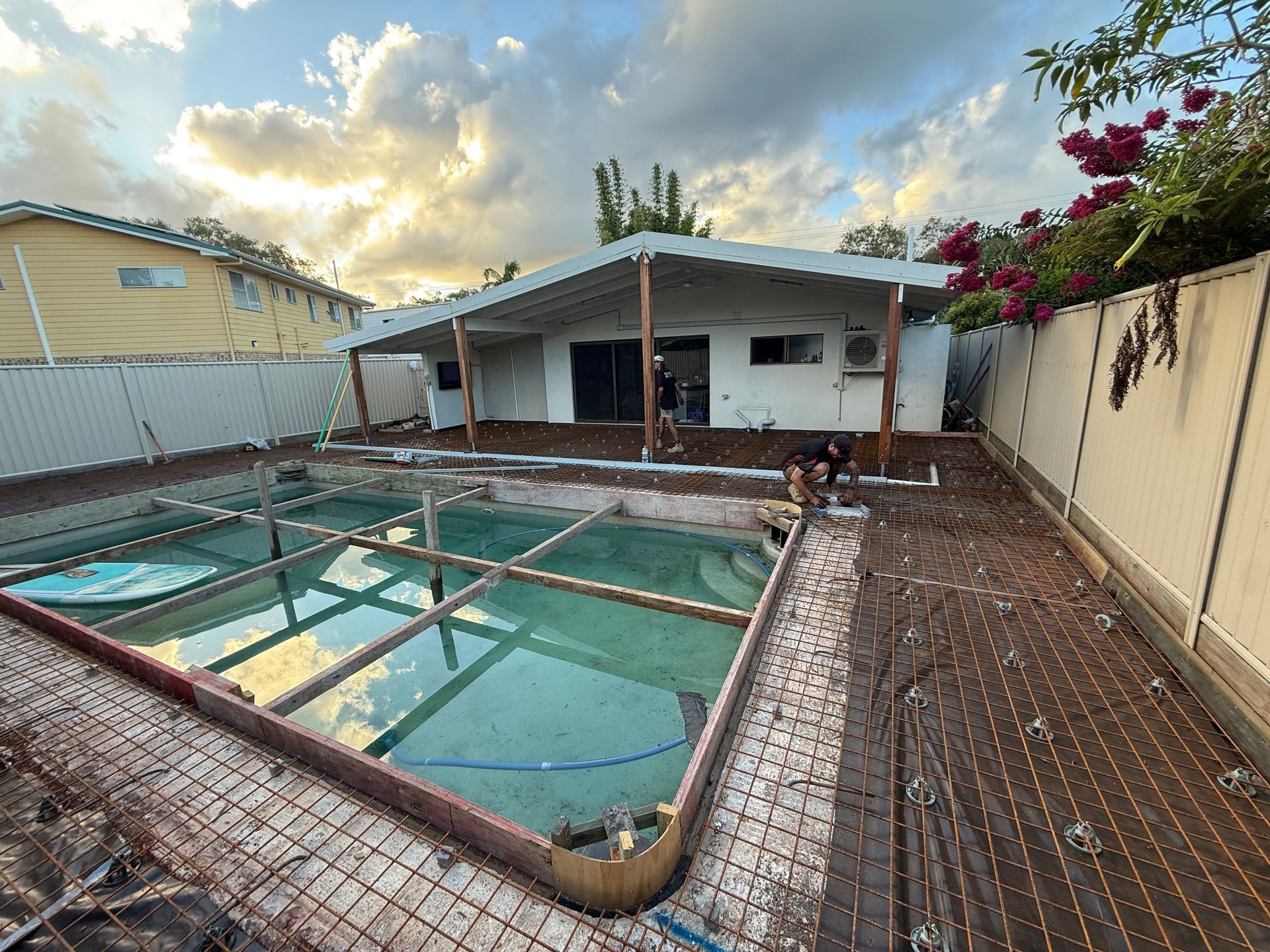 Backyard pool undergoing renovation, with exposed rebar and a partially-finished concrete deck. House in background.