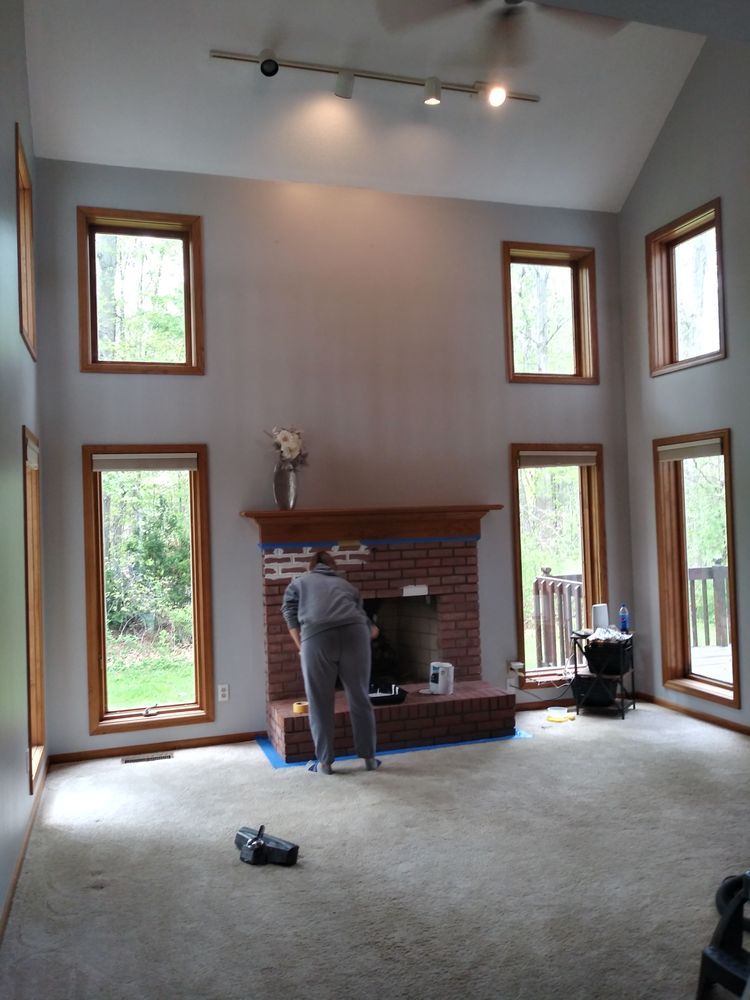 A person paints a brick fireplace in a large living room with tall windows and a high ceiling.