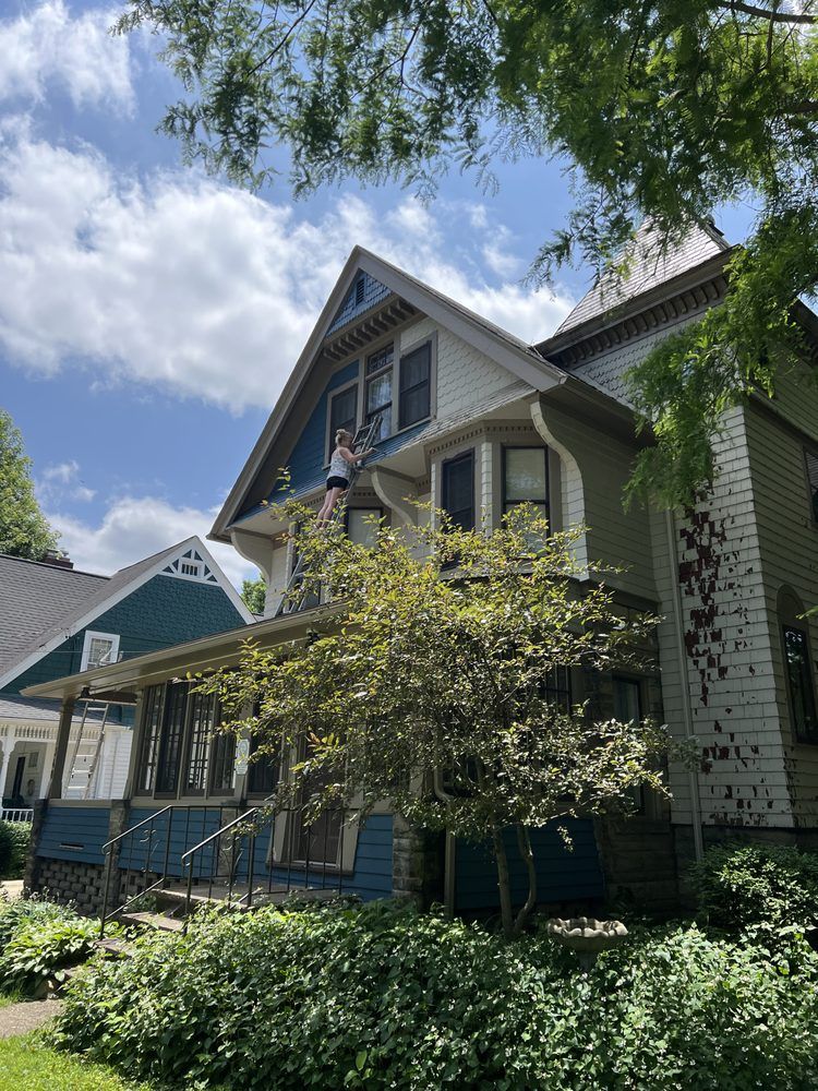 Person on ladder painting a two-story house with blue and white trim. Sunny day. Green foliage in front.