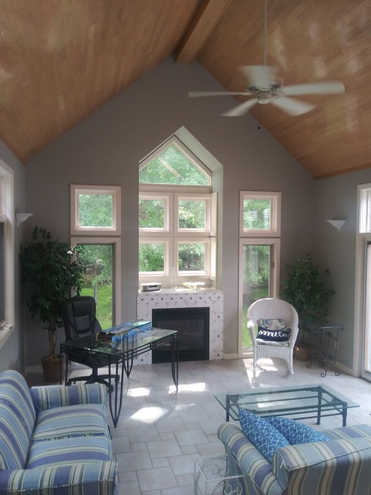 Sunroom with gray walls, wooden ceiling, fireplace, windows, furniture, and plants.