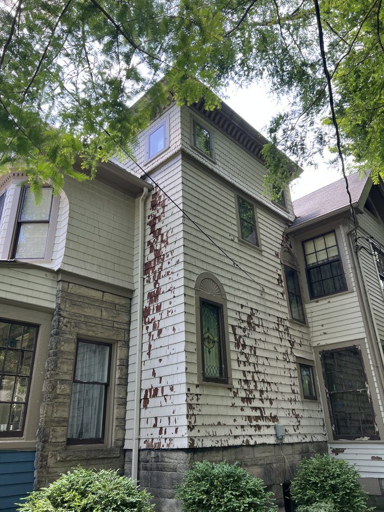 Side of a Victorian house with peeling white paint and various windows, viewed from the ground up.