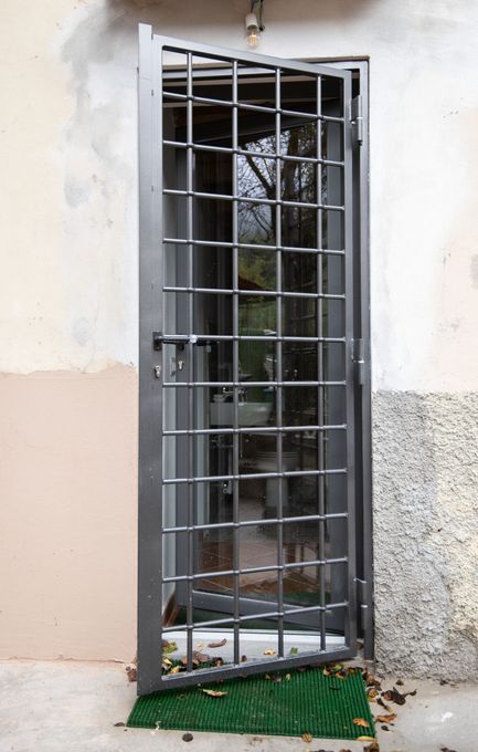 Porta di sicurezza in metallo, socchiusa, con un motivo a griglia sopra una finestra. La porta è montata sulla parete esterna di un edificio.