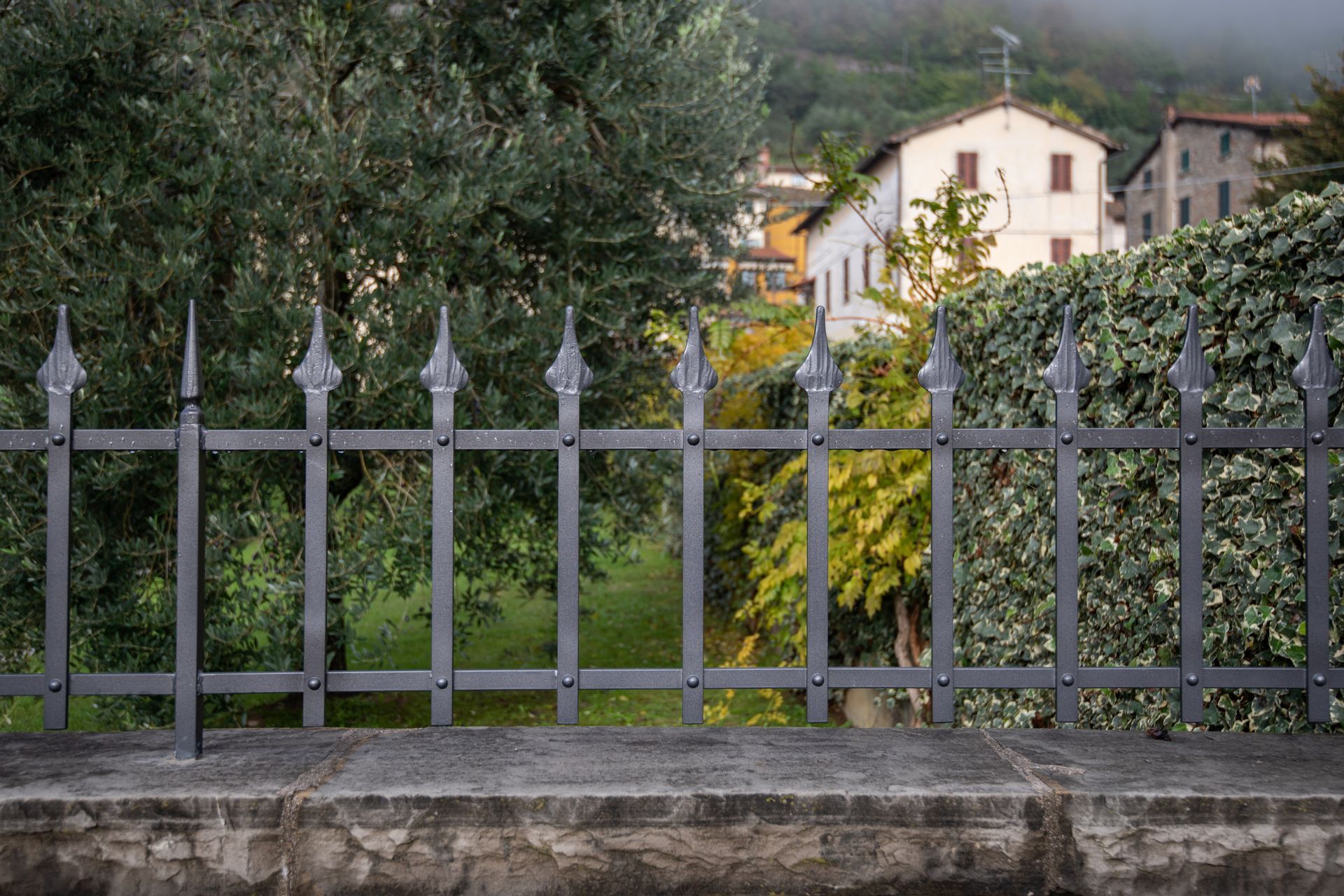 Recinzione in ferro battuto nero con punte decorative, di fronte a un muro di pietra, alberi ed edifici.