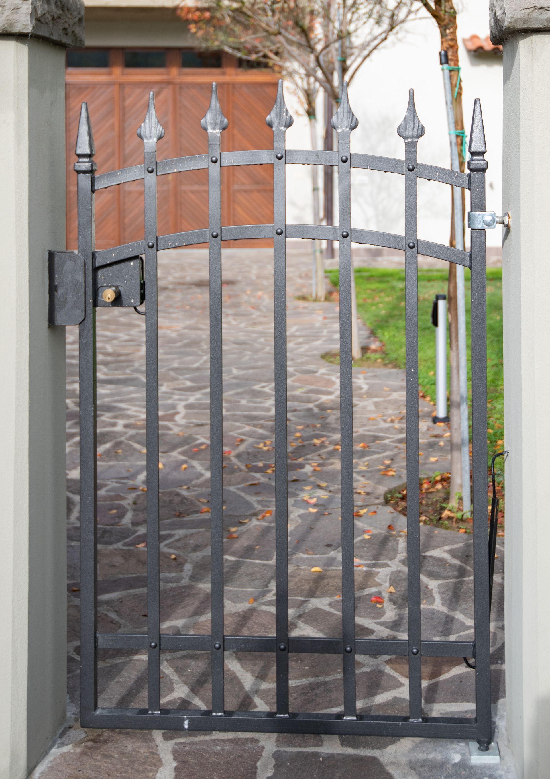 Cancello in ferro nero su un sentiero in pietra, che si apre su un cortile con erba e una porta di legno.