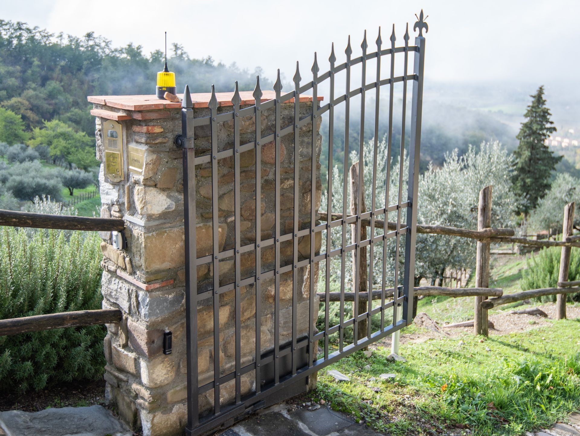 Pilastro in pietra con cancello in ferro battuto, aperto. Recinzione in legno e alberi sullo sfondo.