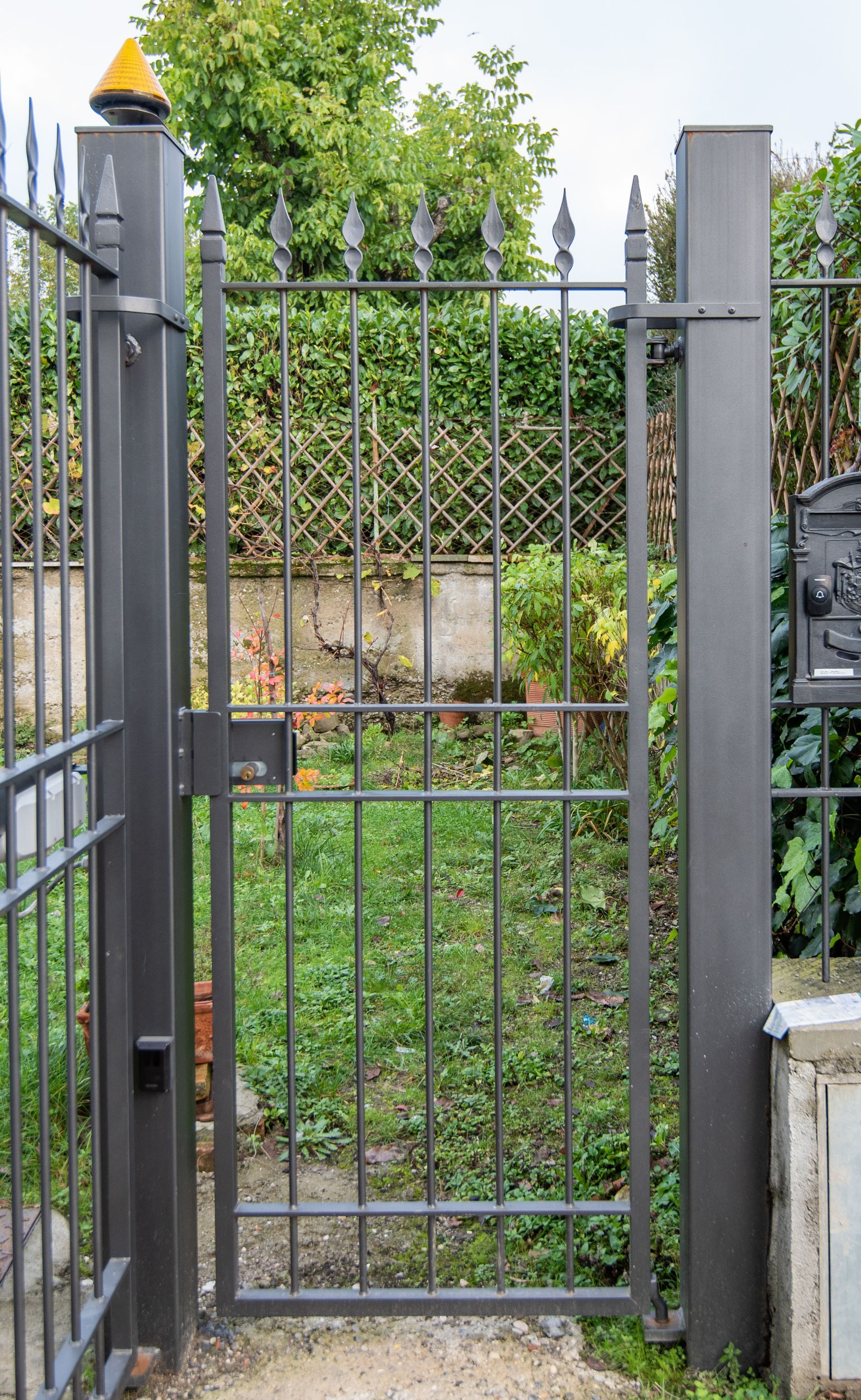 Cancello in metallo grigio scuro, aperto, che conduce a un giardino con erba verde e arbusti.