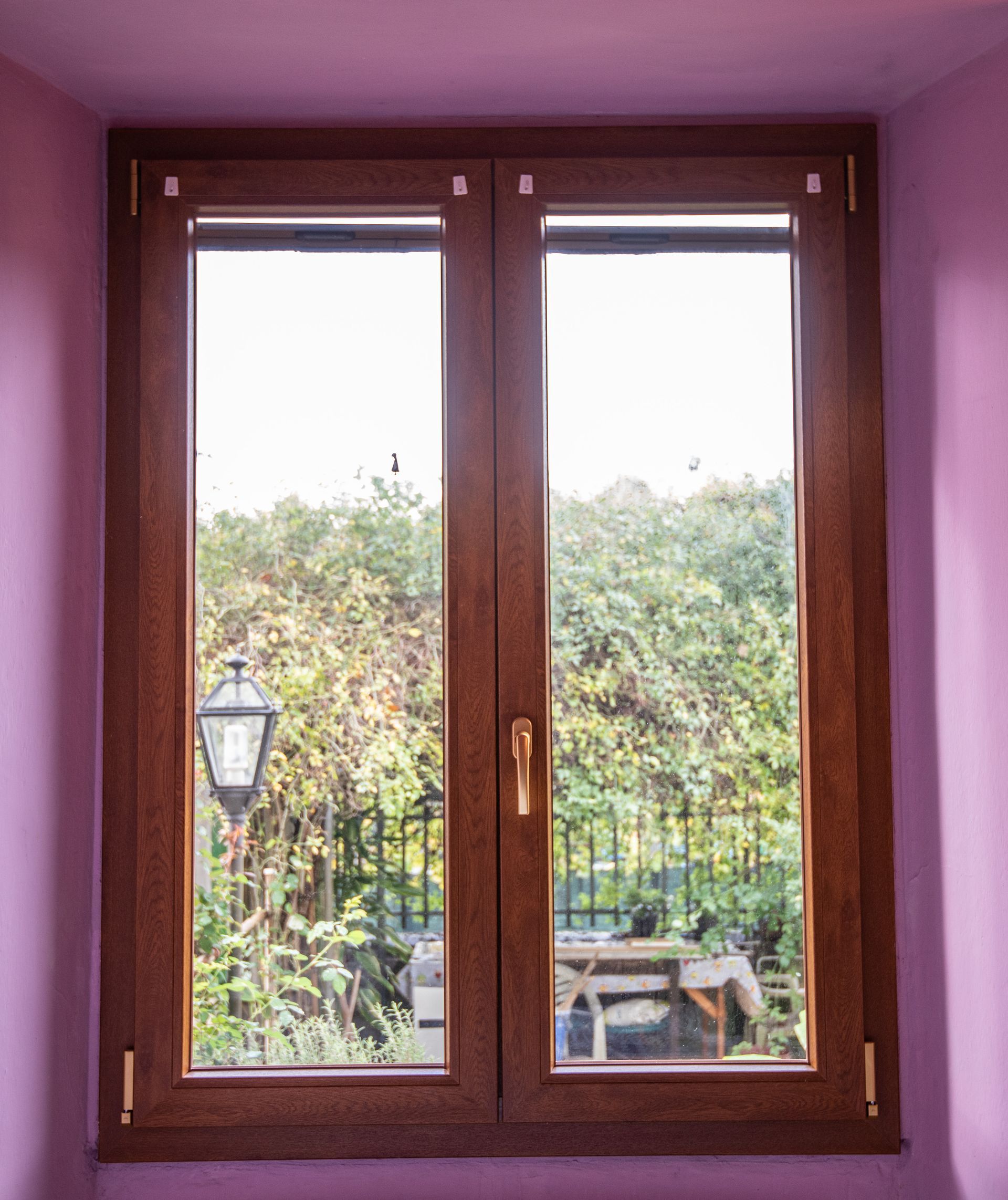 Doppia finestra con cornice in legno, vista sul verde, un lampione e un tavolo. Situata in una stanza con pareti rosa.