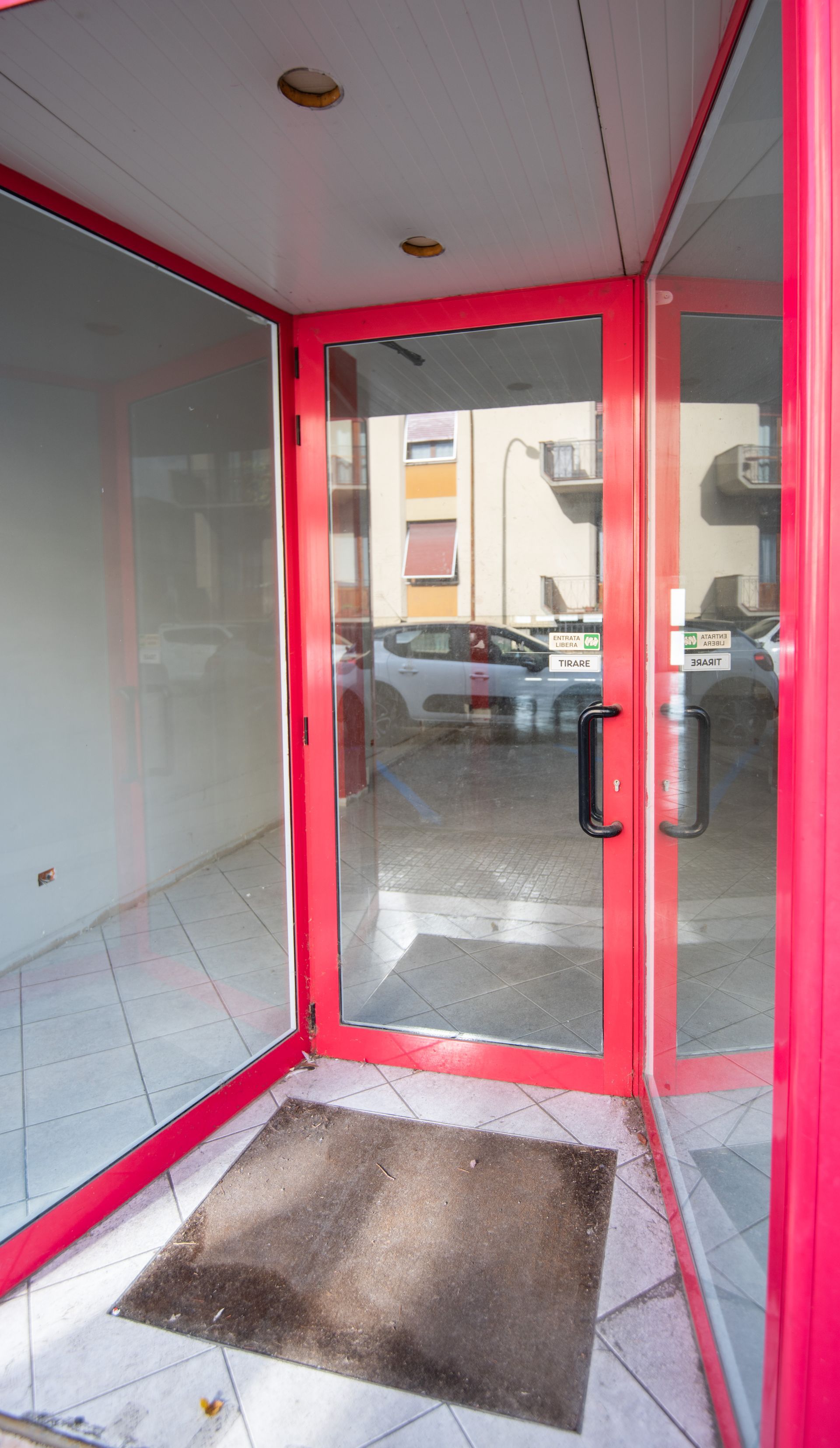 Porta a vetri con cornice rossa e zerbino che conduce a un negozio. All'esterno si vedono un'auto e un edificio.