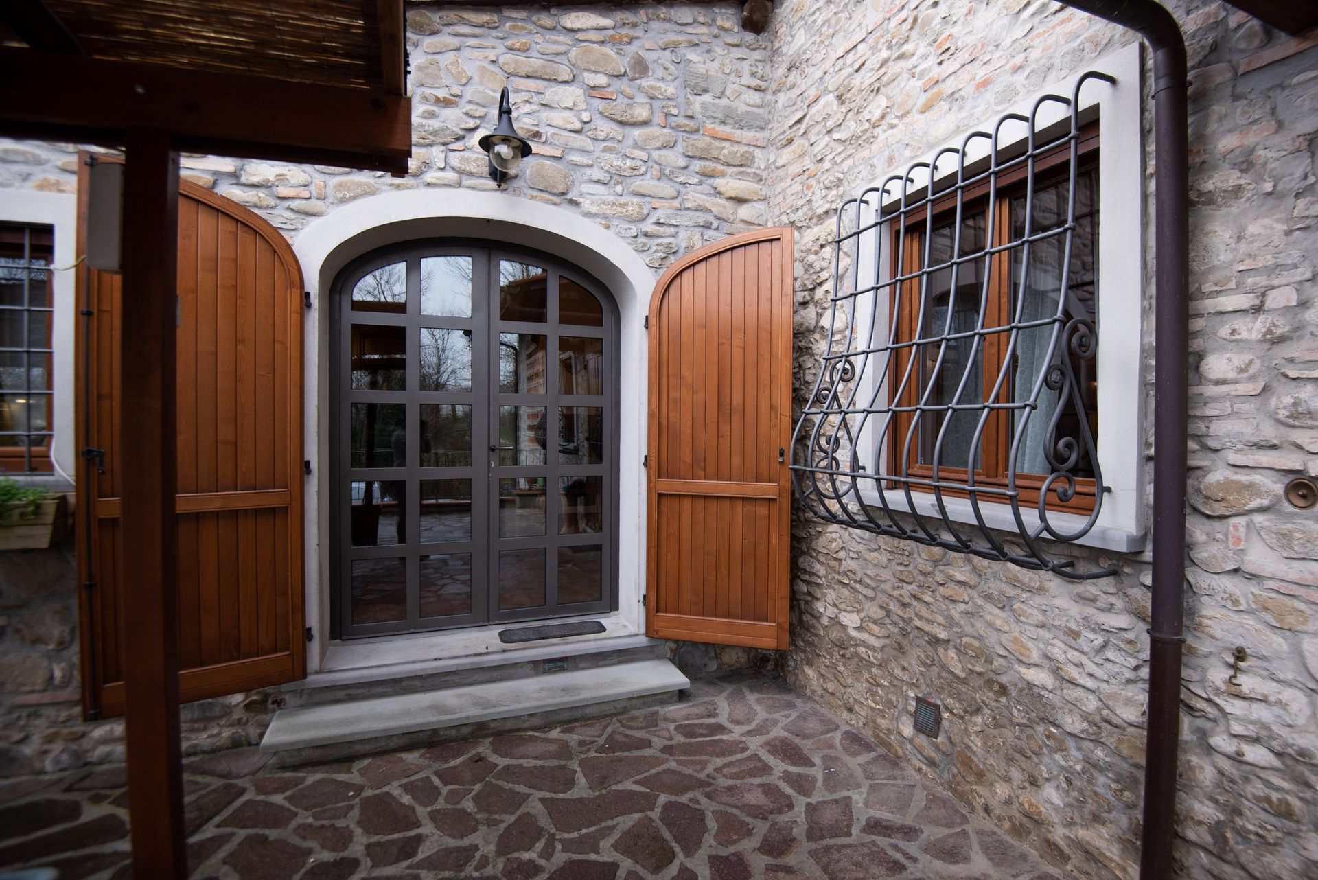 Esterno dell'edificio in pietra con persiane in legno marrone, porta ad arco in vetro e finestra con sbarre.