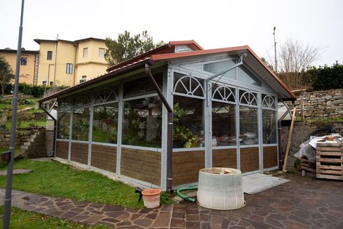 Serra in vetro e legno con tetto rosso, situata accanto a una casa e a un giardino.