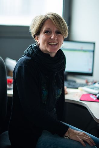 Una donna con i capelli biondi corti sorride, indossa un maglione nero e una sciarpa ed è seduta alla scrivania con un computer.