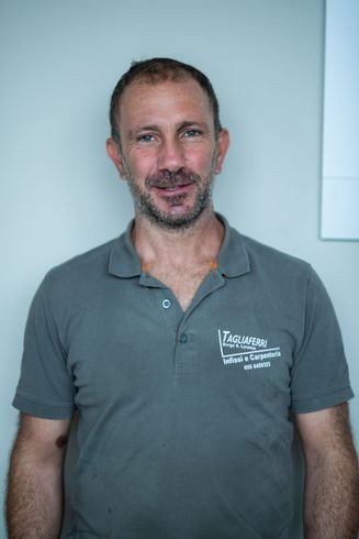 Un uomo con una polo grigia sorride alla telecamera. È in piedi contro un muro grigio chiaro.
