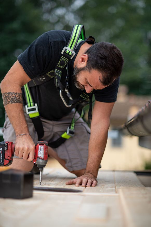Operaio edile inginocchiato, che usa un trapano su una superficie di legno, indossando un'imbracatura di sicurezza.