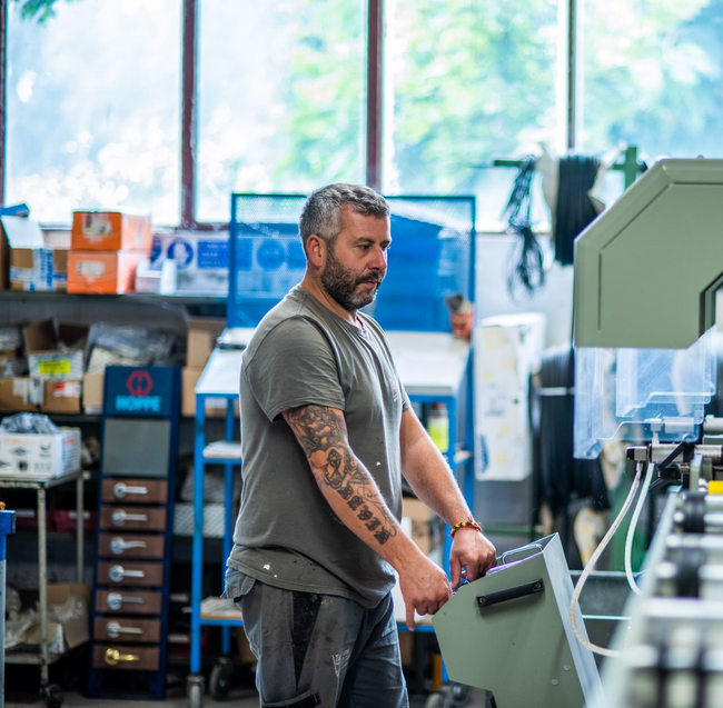 Uomo che aziona macchinari in un'officina, con lo sguardo rivolto verso il basso. Ha tatuaggi e indossa una maglietta grigia e jeans.