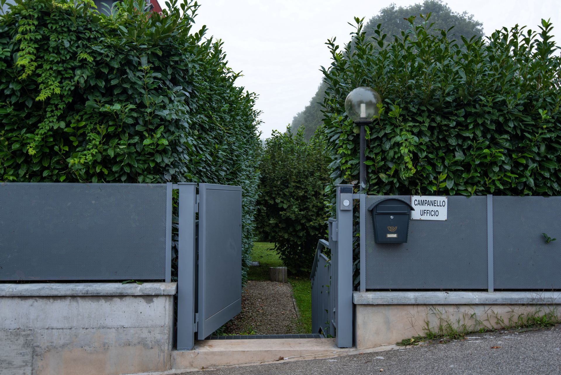 Cancello grigio e recinzione che conducono a un sentiero delimitato da siepi verdi; cassetta postale e lampione sulla destra.