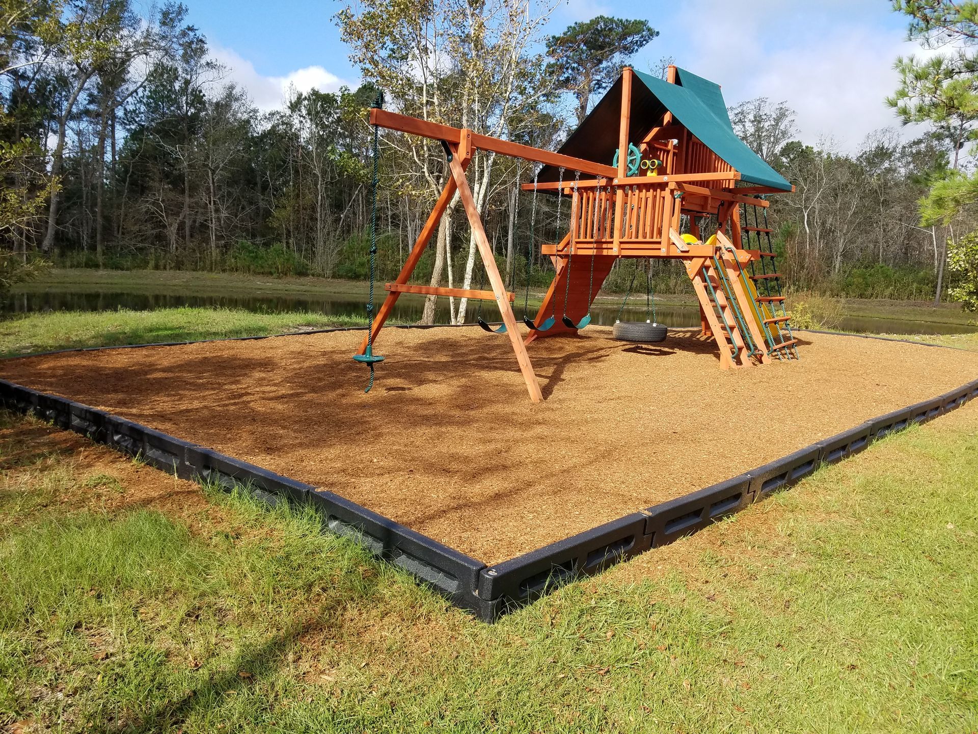 Wooden playset with swing set and slide, on woodchip base, bordered by black edging, set in grassy area.