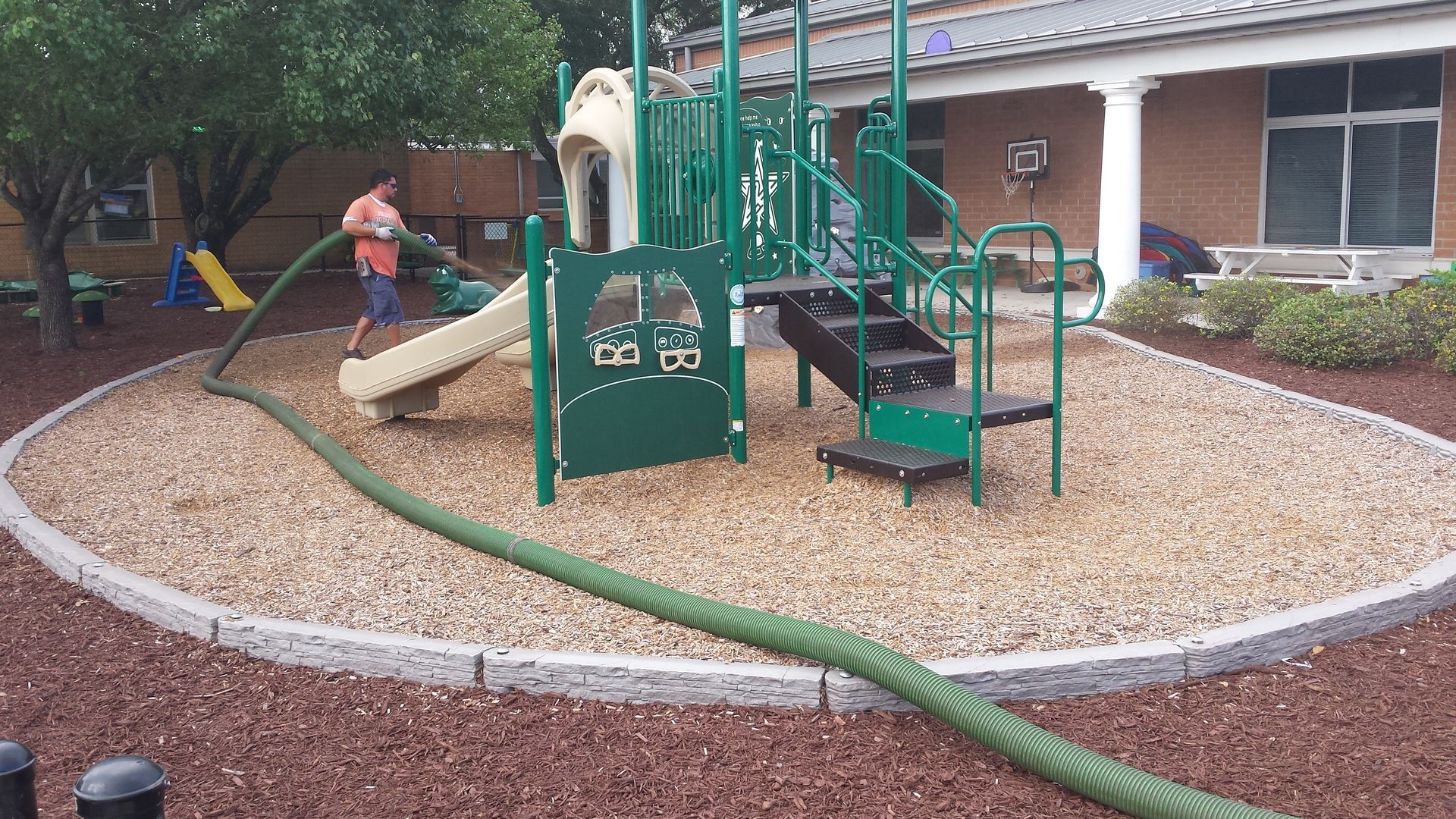 Person spraying mulch around a green playground structure with slides and stairs, near a building.