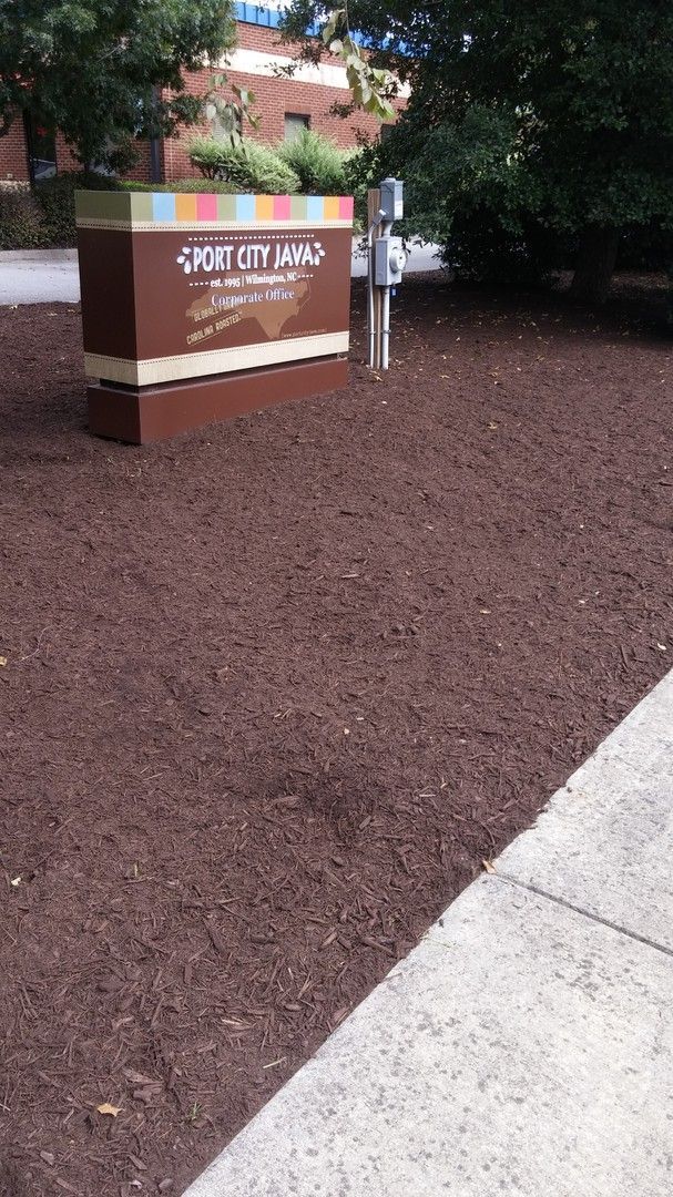 Sign for Sweet Lily Lane in a mulched area, with a building and sidewalk visible.