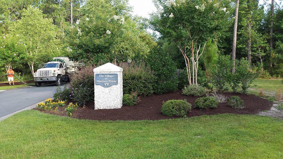 Entrance sign with landscaping; truck unloading. Green grass, trees, and shrubbery.