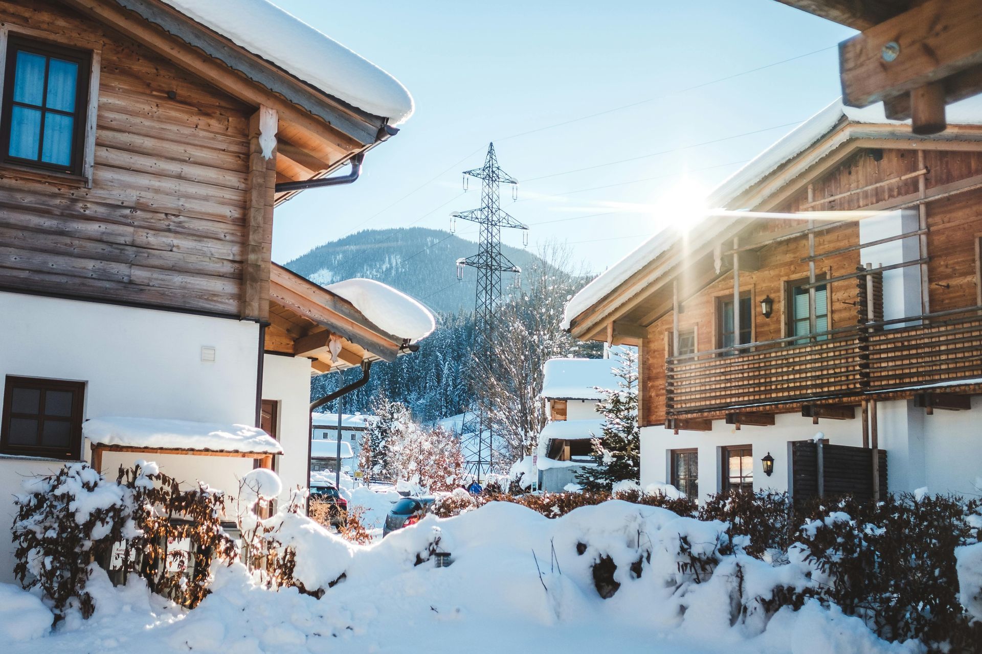 Snow-covered wooden chalets bask in winter sun, with a mountain and power lines in the background.