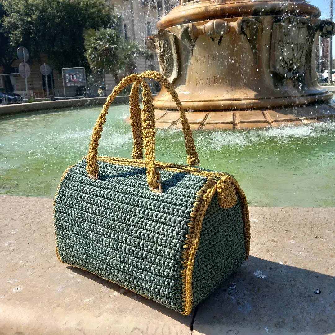Borsa a mano verde intrecciata con manici dorati su una sporgenza di pietra accanto a una fontana all'aperto.