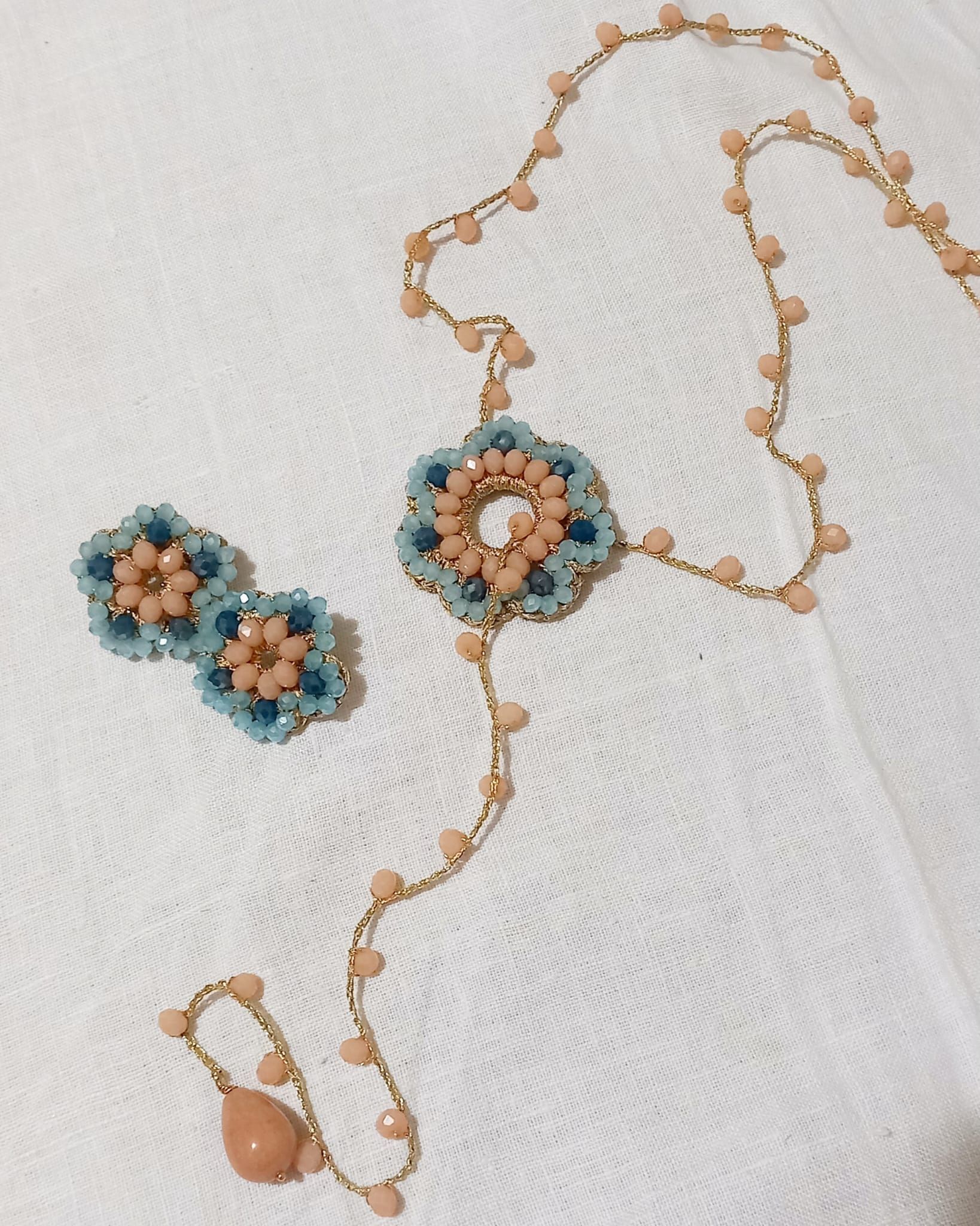 Set di gioielli con perline composto da collana floreale turchese e color pesca, orecchini coordinati e anello su tessuto bianco.
