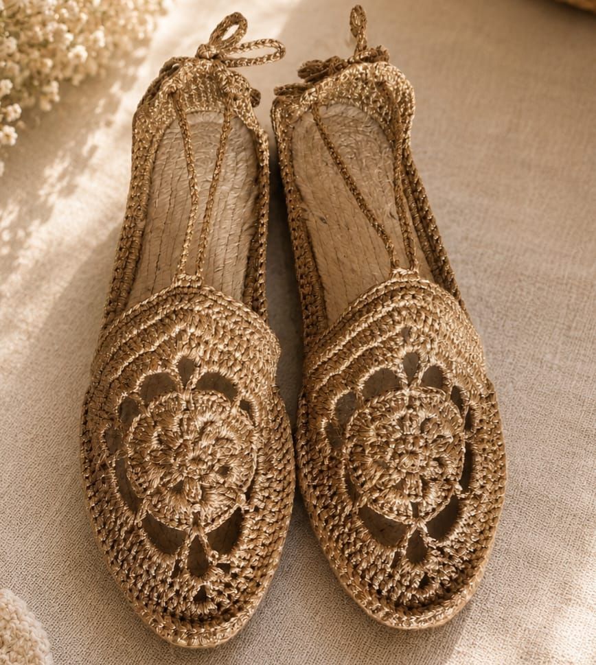 Un paio di ballerine slip-on in maglia dorata con dettagli floreali intagliati su una superficie in tessuto beige.