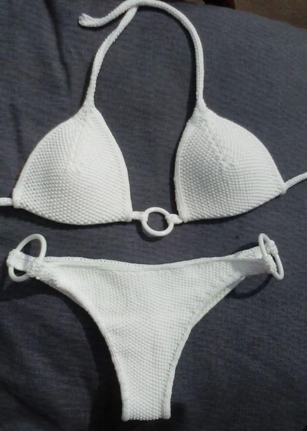 Bikini bianco testurizzato con top a triangolo e slip con laccetti laterali su sfondo di tessuto scuro.