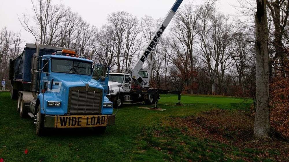 Blue crane truck lifting a long pole in a grassy, tree-lined area with a yellow “WIDE LOAD” sign.