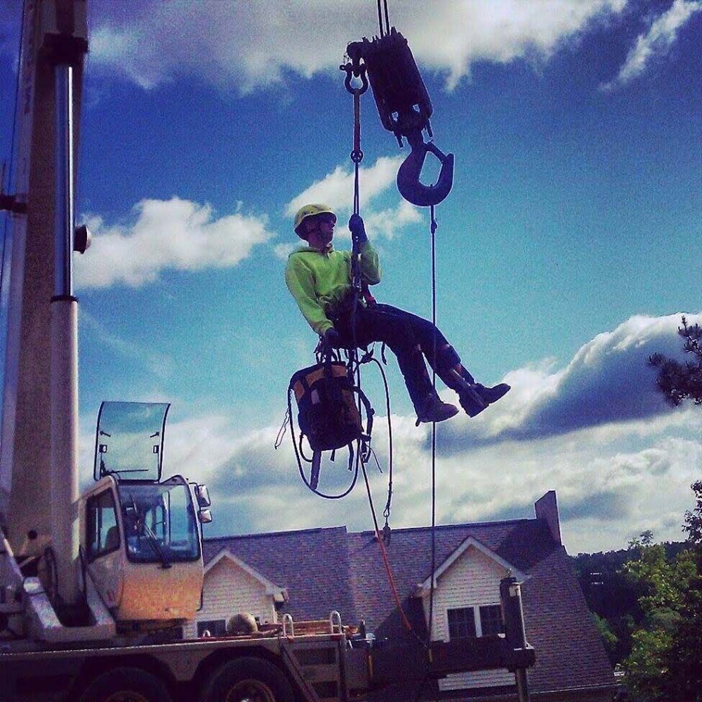 Construction worker suspended from a crane, wearing a helmet and safety harness, against a blue sky.