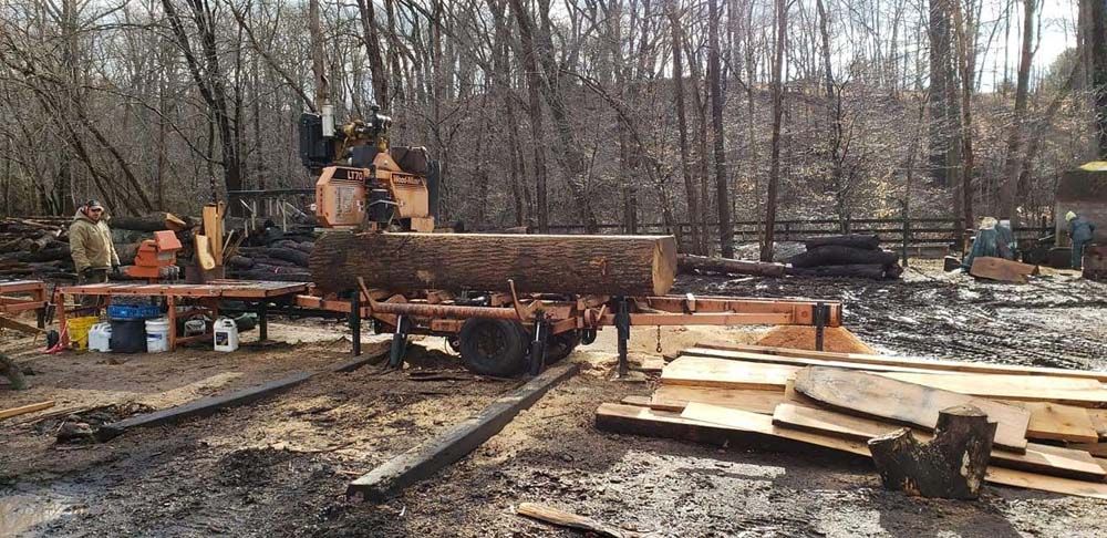 Excavator on a muddy construction site with lumber piles and bare trees in the background