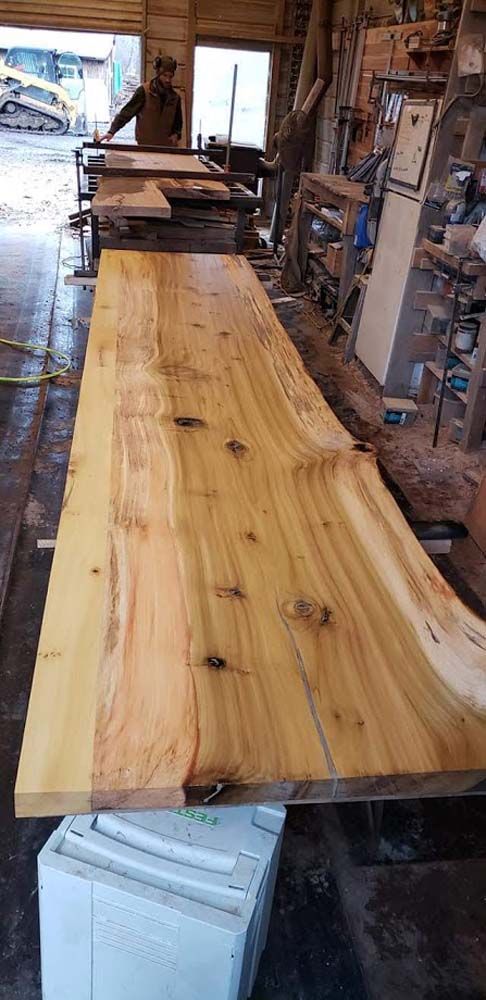 Long live-edge wooden slab on supports in a rustic workshop, with a person working in the background