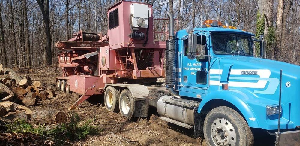 Blue semi truck hauling a red wood chipper in a wooded clearing with cut logs nearby