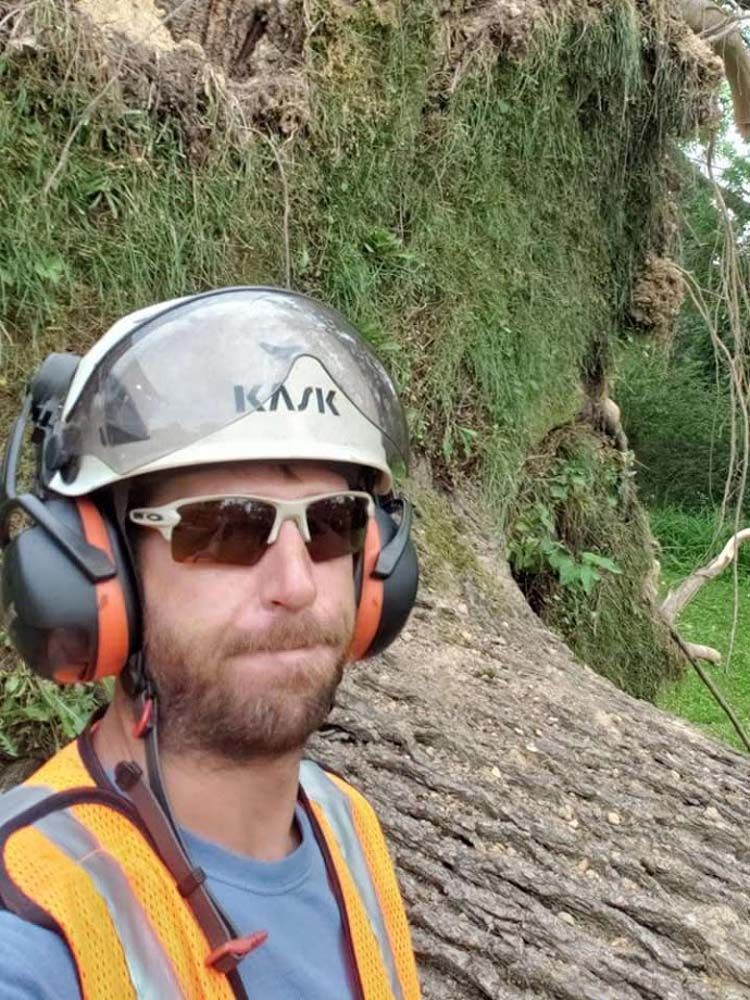 Worker in hard hat and ear защитors standing by a tree trunk and grassy embankment.