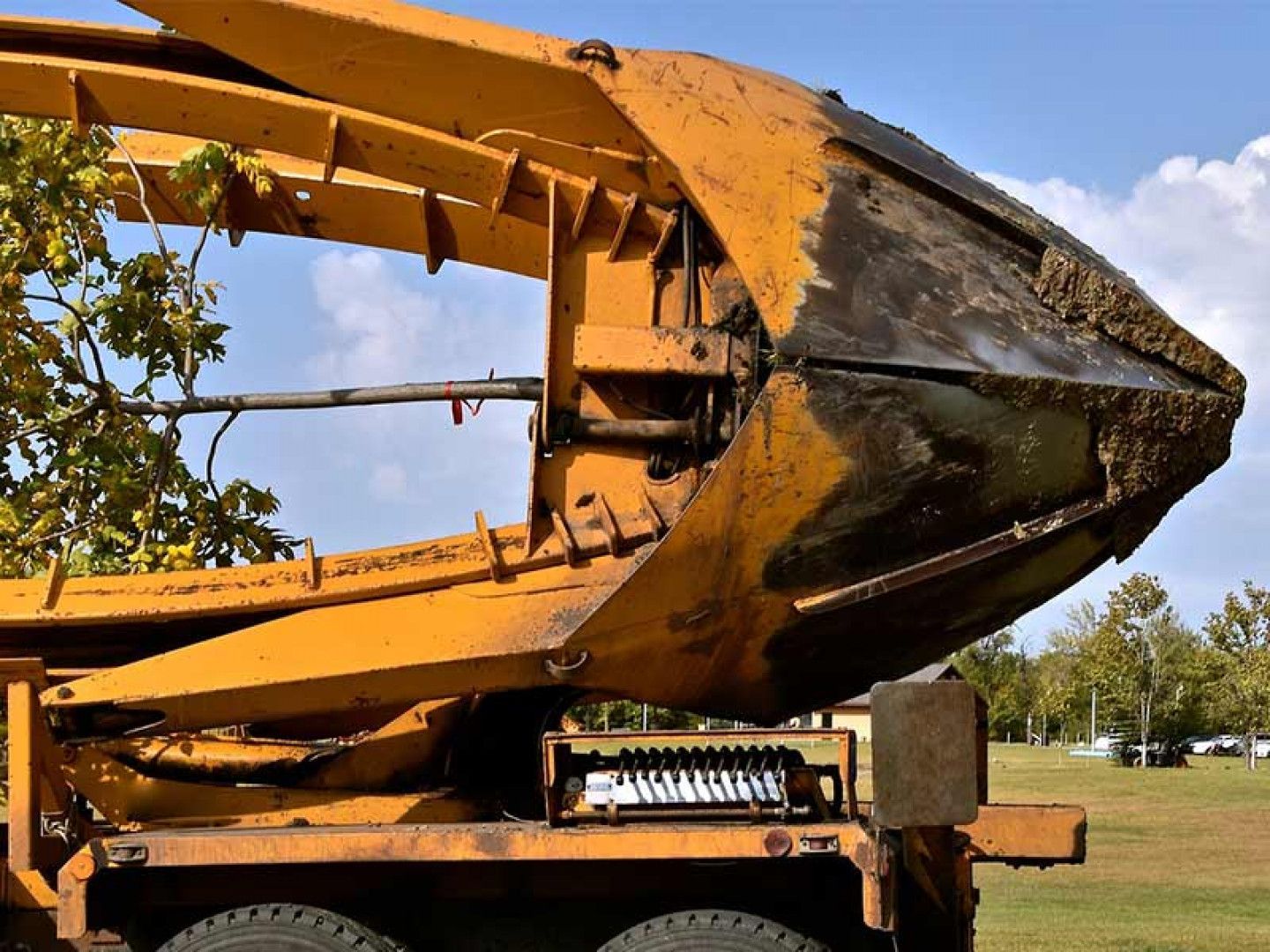 Rusty yellow excavator bucket on a trailer in a grassy field under a blue sky