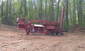 Red industrial wood processor beside a dirt mound in a wooded clearing, with a person standing nearby.