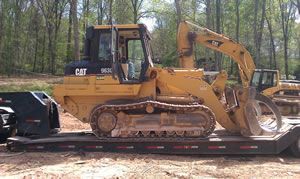 Yellow CAT excavator loaded on a flatbed trailer in a wooded outdoor area.