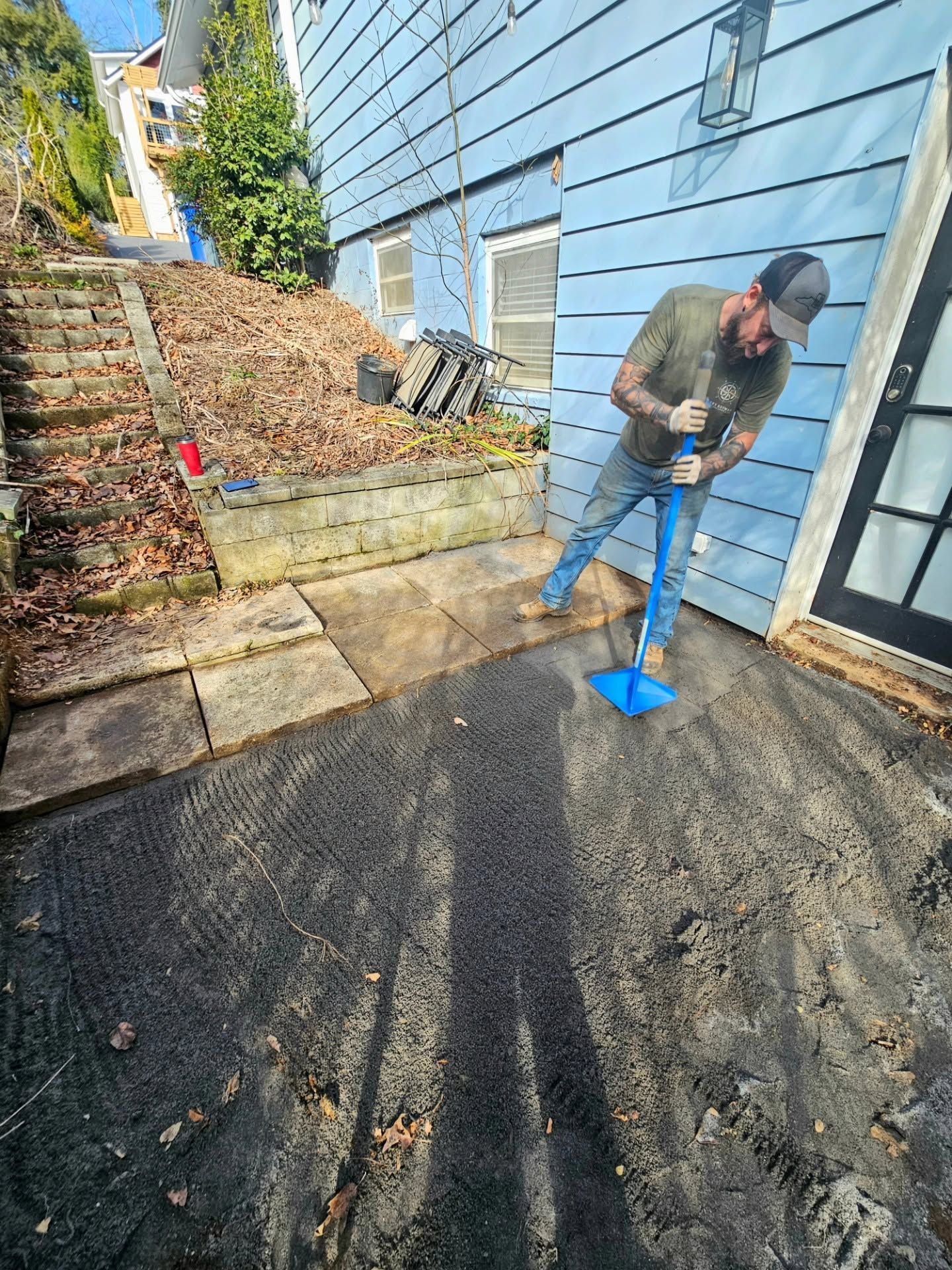 Man shoveling wet dirt from a driveway beside a blue house, with paving stones and a compost pile nearby