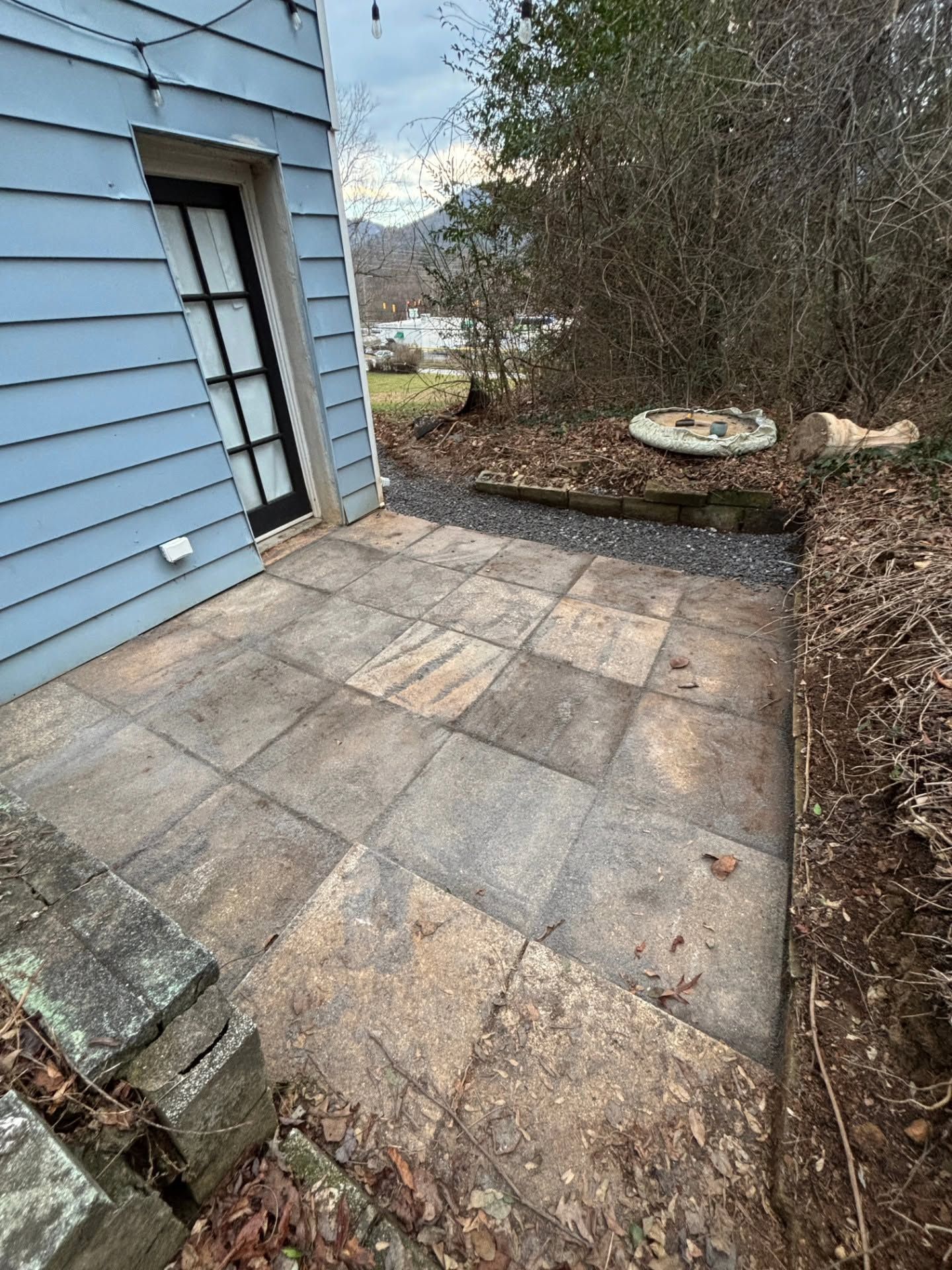 Stone patio beside blue house with a doorway, bordered by mulch and bare trees.