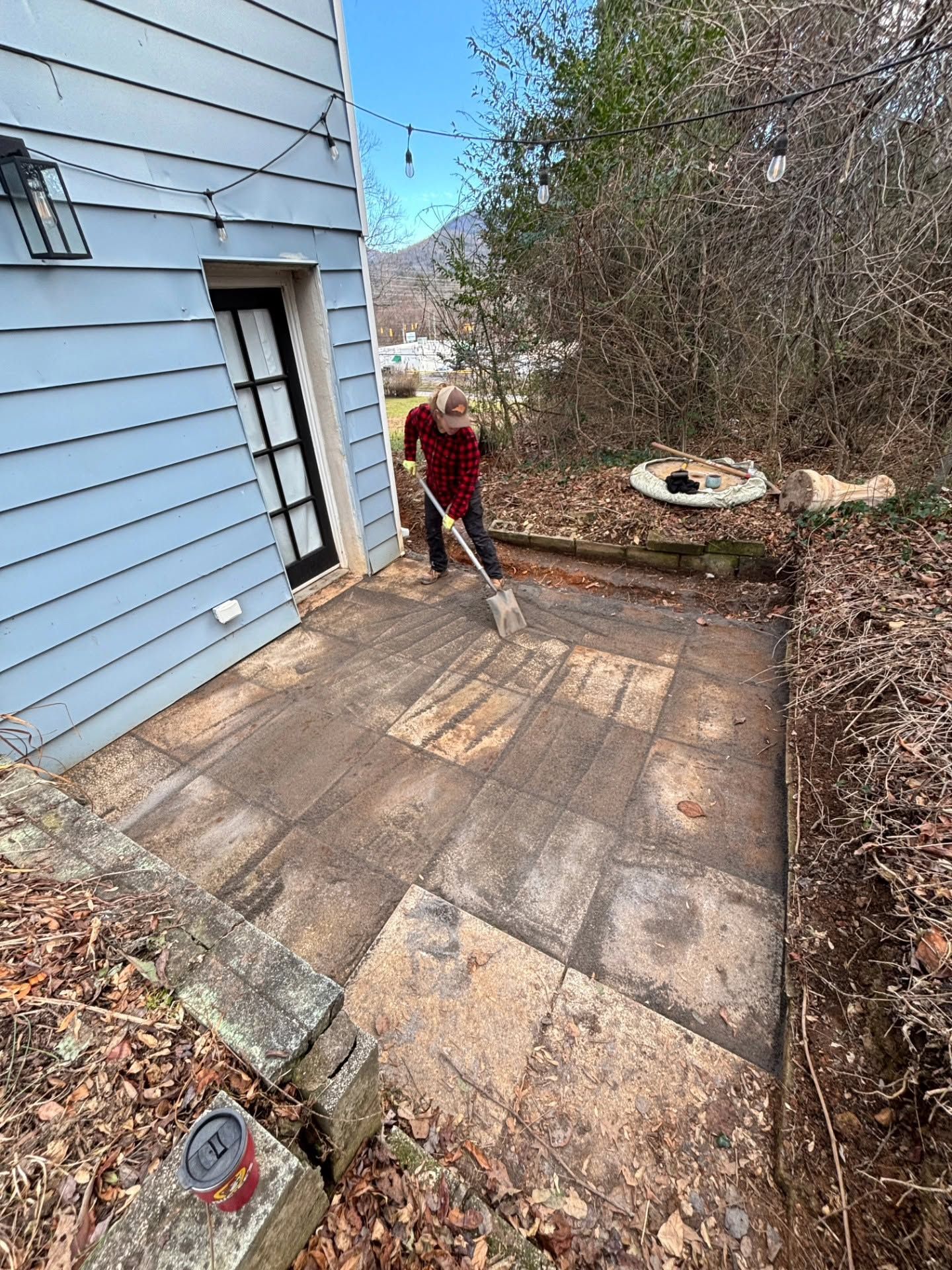 Person sweeping a stone patio beside a blue house, with brush and wooded yard nearby