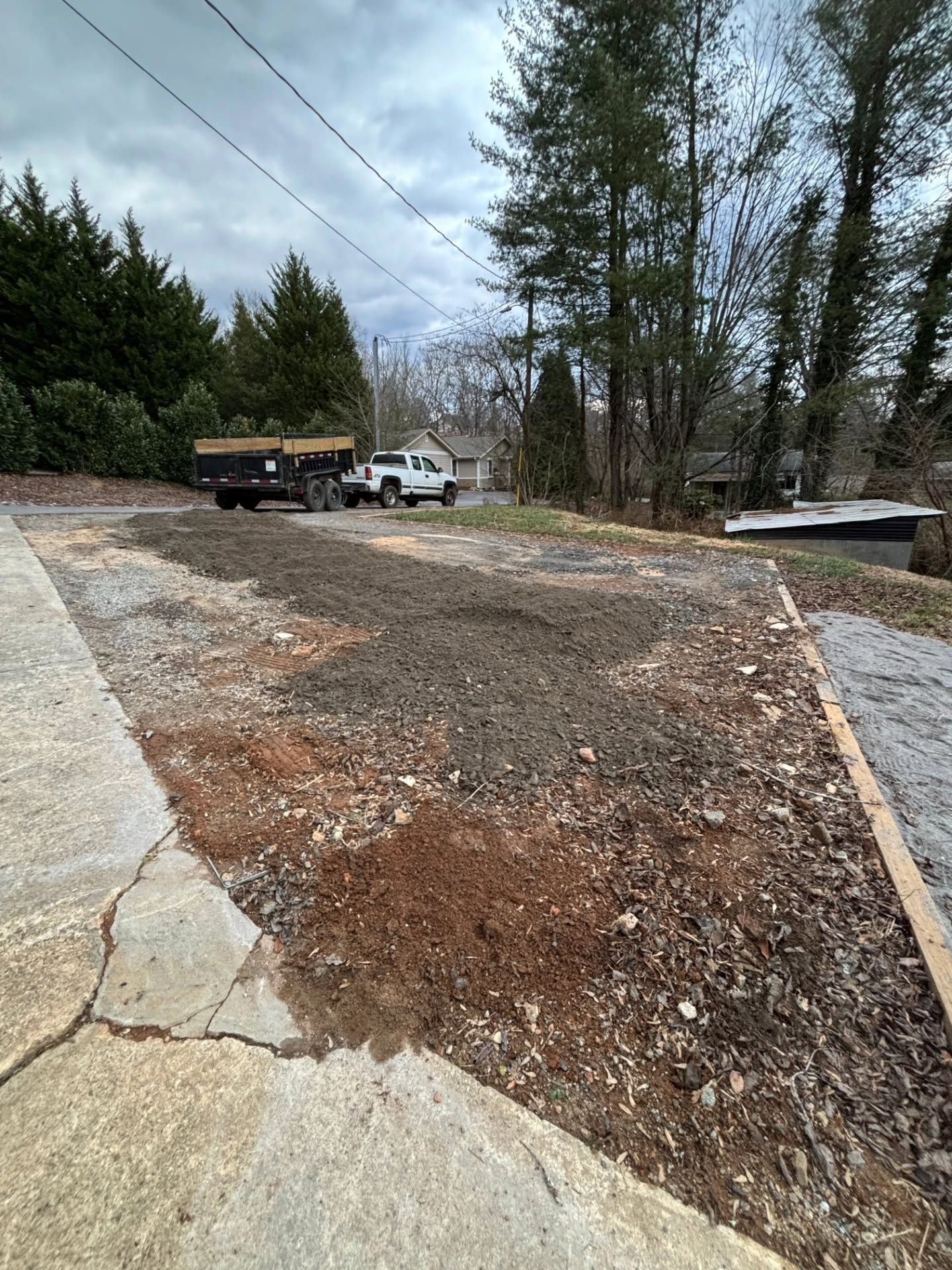 Gravel driveway beside a concrete slab, with two parked trucks and tall trees under a cloudy sky
