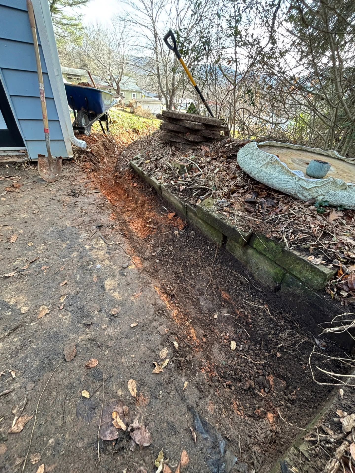 Narrow dirt path beside a house, with a raised garden bed and trees in a wooded yard.