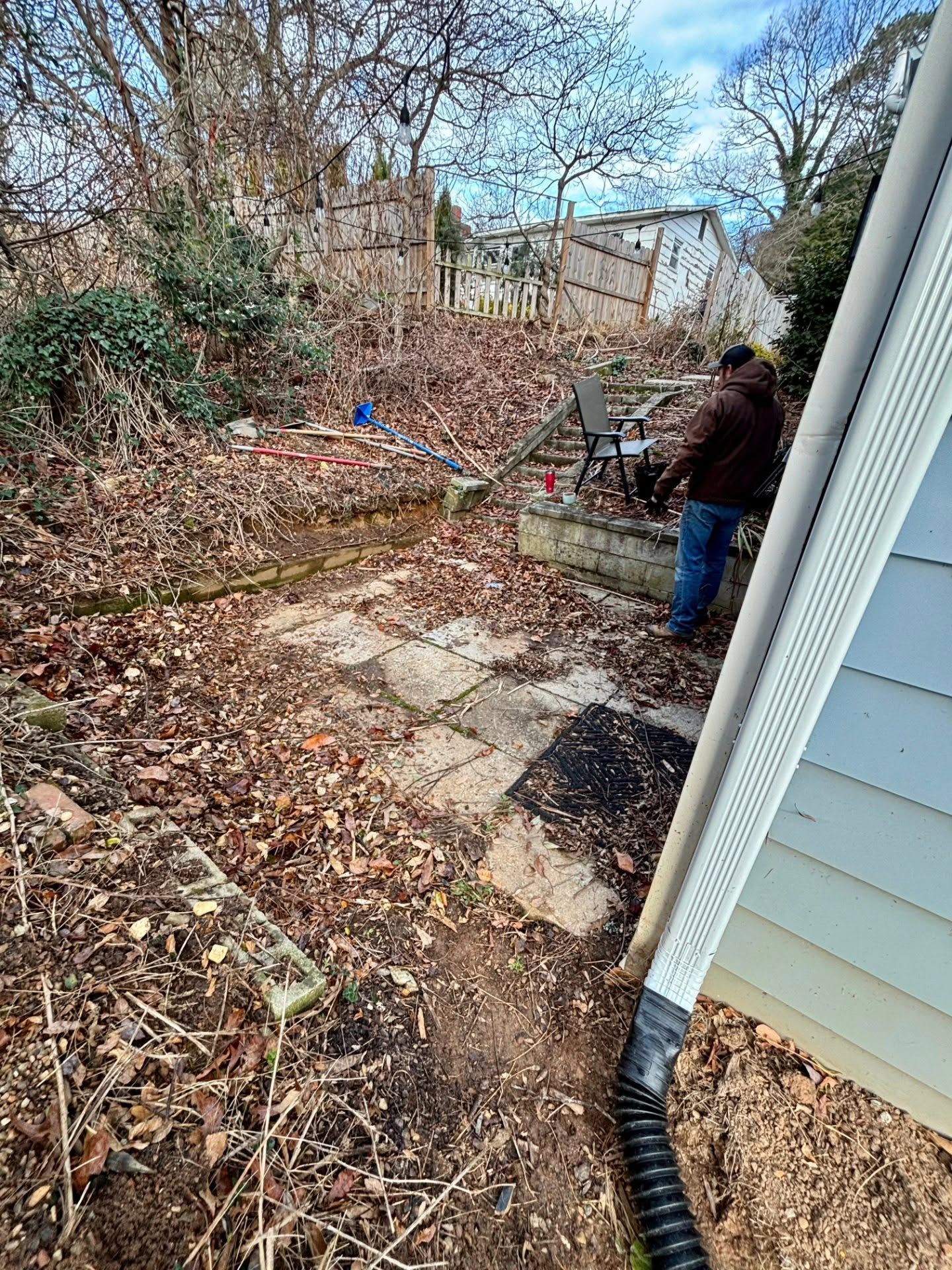 Person standing in a leaf-covered backyard beside a house and a raised stone patio.