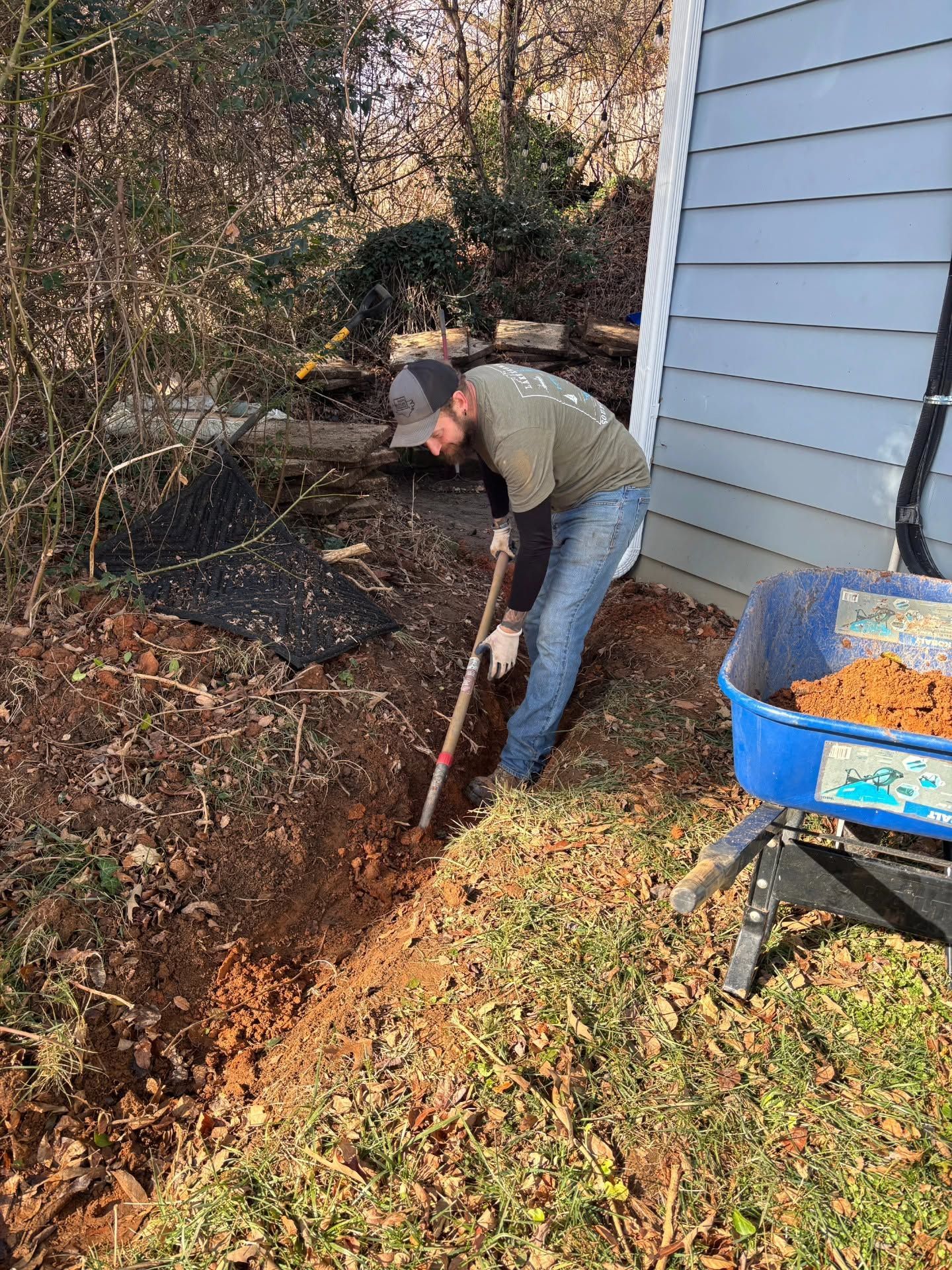 Person digging in a leaf-covered yard beside a house with a shovel and blue wheelbarrow.