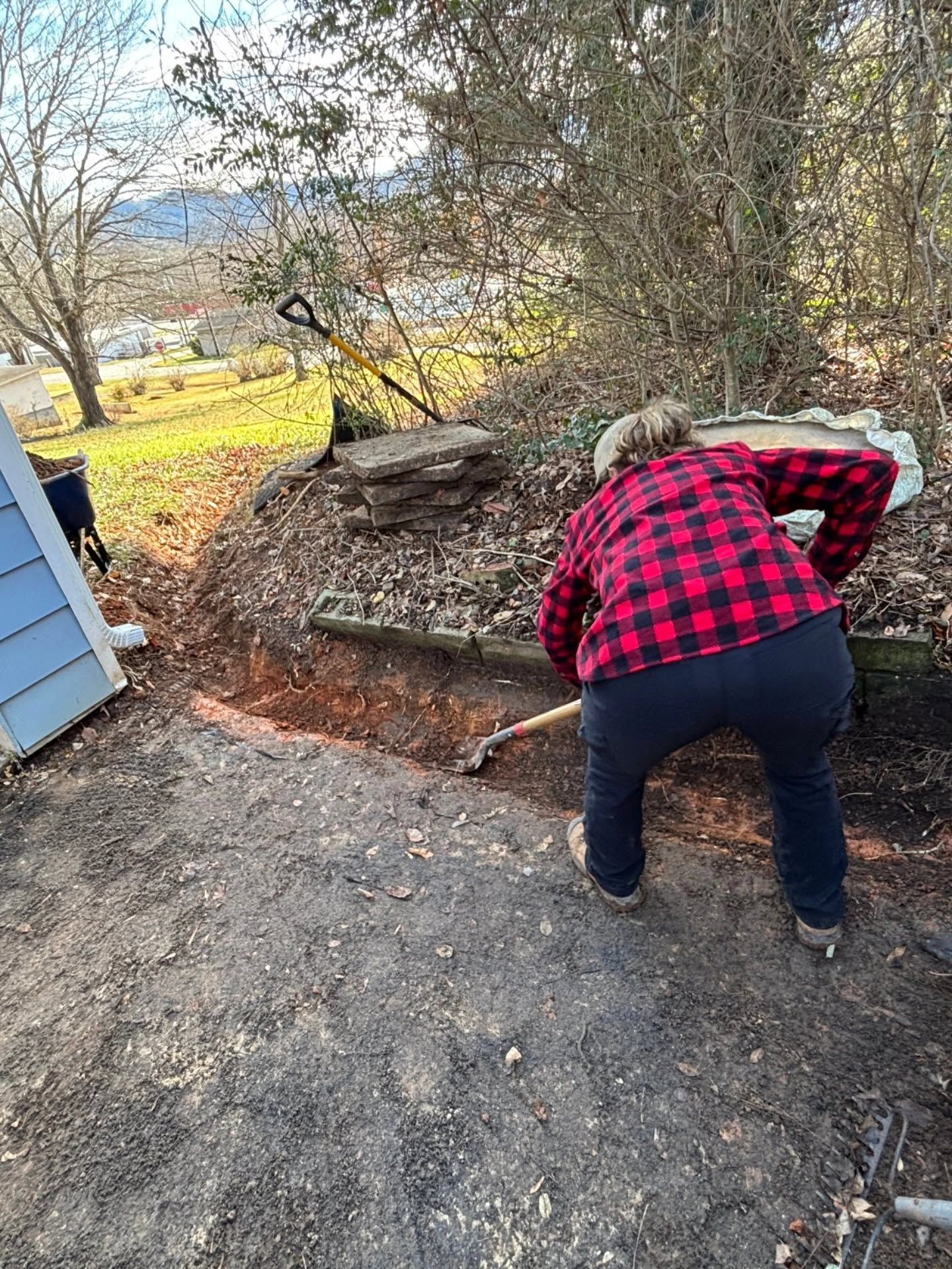 Person in red plaid shirt digging along a garden edge beside a blue building and stone pile.