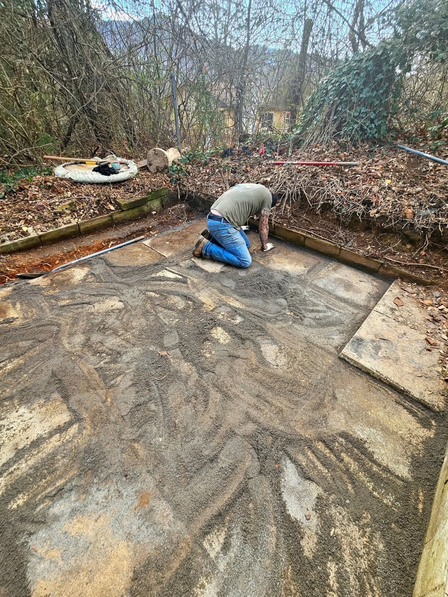 Person kneeling on a stone patio, scraping mud or debris from the ground in a garden area.